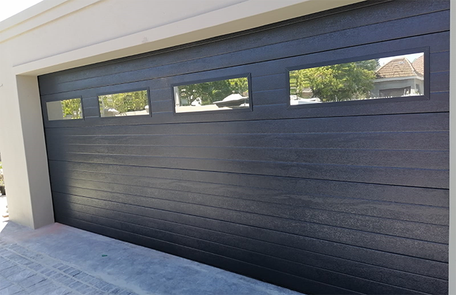 Modern black garage door with four windows, featuring a sleek design in a residential setting.