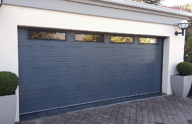 Modern blue garage door with windows, flanked by potted plants, on a paved driveway.