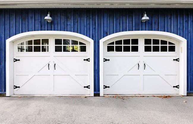 White double garage doors on a blue siding wall, featuring arched windows and exterior lighting above.