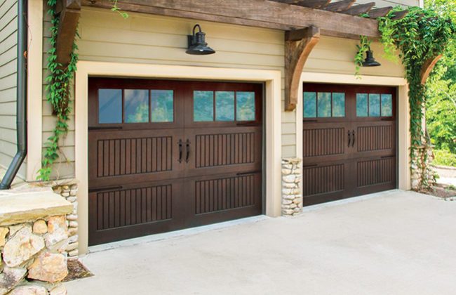 Two elegant wooden garage doors with glass panels, framed by stone accents, under a pergola with lush greenery.
