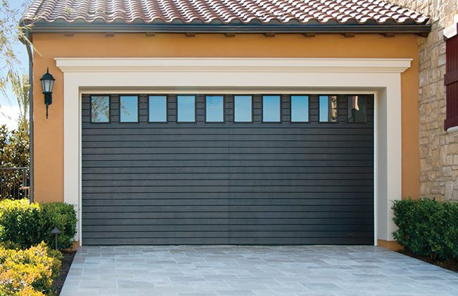 Modern brown garage door with small windows, bordered by beige trim and surrounded by a tiled driveway and green shrubs.