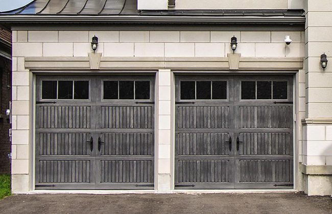 Two gray wooden garage doors with decorative windows and outdoor lighting on a residential building facade.
