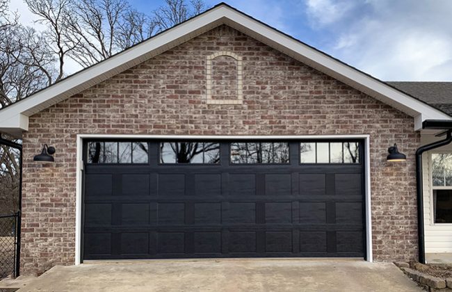 Modern brick garage exterior with black door and outdoor lighting fixtures under a clear blue sky.