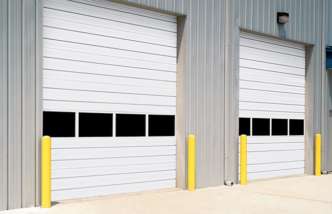 White industrial garage doors with windows and yellow bollards on a concrete pad.