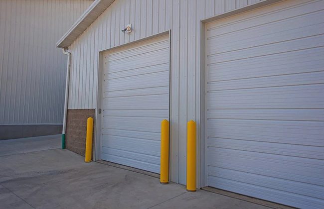 White industrial building with two metal garage doors and yellow bollards under a clear sky.