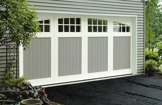 Modern house with three-panel garage door, white frame, glass windows, and gray siding surrounded by greenery.