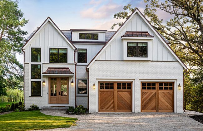 Modern white farmhouse with wooden garage doors and large windows surrounded by trees.